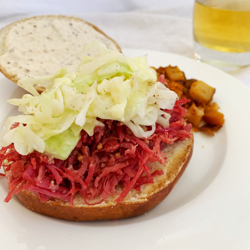 A picture of a pulled corned beef and cabbage sandwich open-faced as it's being assembled with potatoes in the background. 