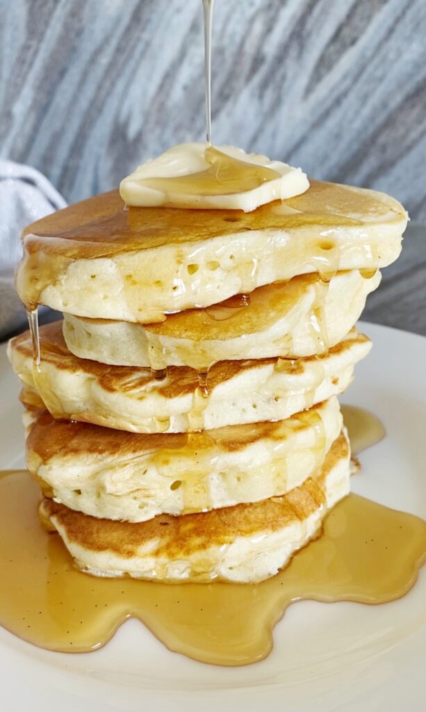 A stack of puffy pancakes topped with a pat of butter being drizzled with warm maple syrup.