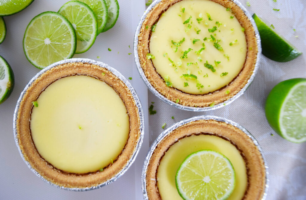 A picture of lime and goat cheese individual cheesecakes sitting on a cutting board - one plain, one with zest, and the last with a slice of lime