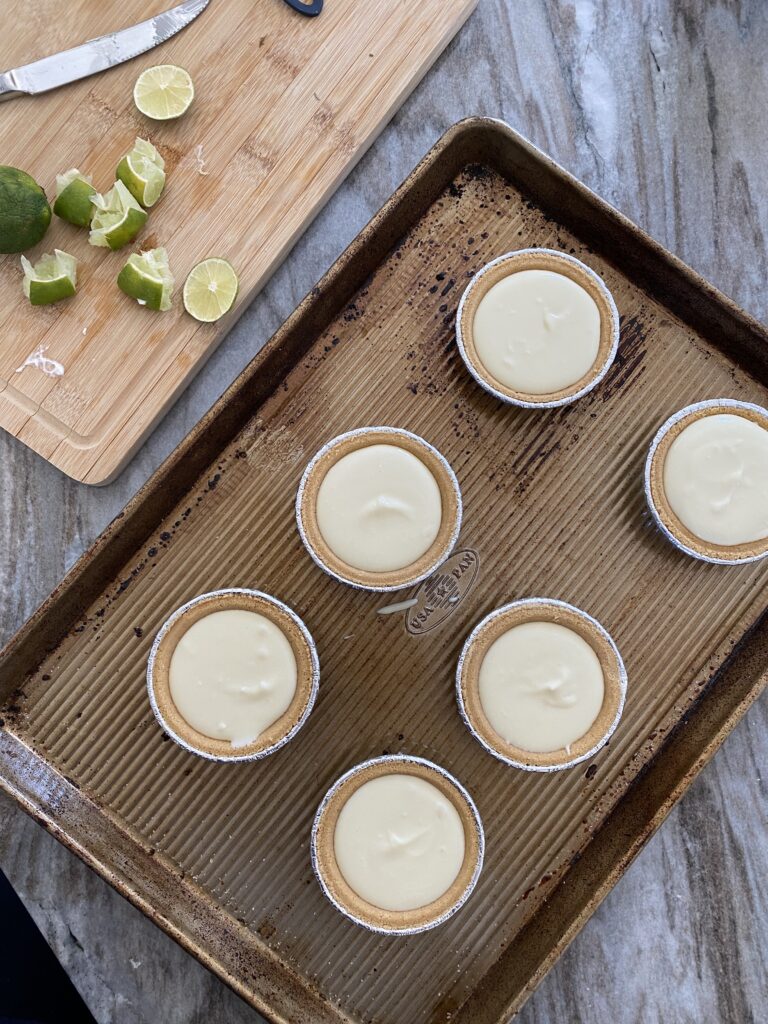 A picture of mini cheesecakes ready for the oven next to a cutting g board with cut up limes