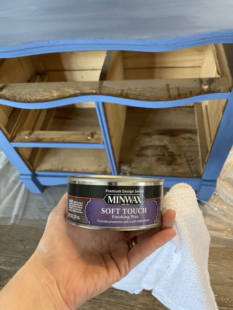 A picture of a hand holding a can of minwax soft touch finishing wax in front of a blue painted dresser.