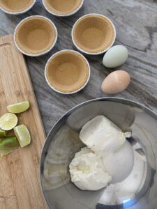A picture of a mixing bowl with cream cheese, goat cheese, and sugar before it's mixed with limes, eggs, and pie shells in the background