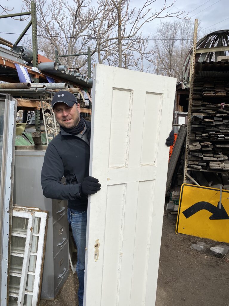A picture of a man holding a white door in the middle of a salvage yard